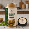 A jar of SSRYJA Coriander Powder with its lid placed beside it on a kitchen countertop, surrounded by coriander seeds and a sprig of cilantro. The background shows a softly blurred kitchen setting.
