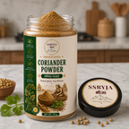 A jar of SSRYJA Coriander Powder with its lid placed beside it on a kitchen countertop, surrounded by coriander seeds and a sprig of cilantro. The background shows a softly blurred kitchen setting.