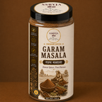 A jar of SSRYJA Garam Masala spice mix on a solid brown background. The jar has a cream-colored label with dark brown text and illustrations of spices, and a black lid with the SSRYJA brand logo.