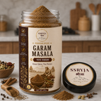 A jar of SSRYJA Garam Masala spice mix with its lid placed beside it on a marble countertop, surrounded by whole spices and a blurred kitchen background.