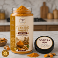 A jar of SSRYJA Turmeric Powder with its lid placed beside it on a marble surface, surrounded by turmeric roots and a small pile of orange powder. The jar has a cream label with gold and brown text, and the lid is black with a matching cream label.