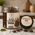 A jar of SSRYJA Black Cardamom premium spices next to its lid, with whole cardamom seeds scattered on a marble surface. The background shows a softly blurred kitchen setting with wooden utensils.