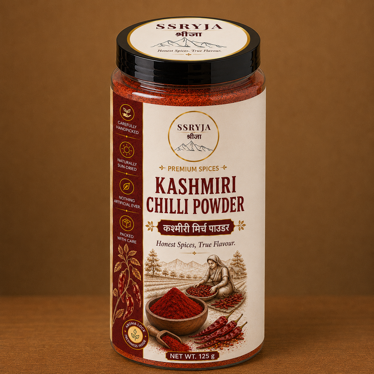 A jar of SSRYJA Kashmiri Chilli Powder on a brown background. The jar has a cream-colored label with red and black text, showing illustrations of chili peppers and a person grinding spices.