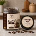 A jar of SSRYJA Clove spices with its lid placed beside it on a marble surface, with a few loose cloves scattered in front. The background shows a softly blurred kitchen setting with wooden containers.